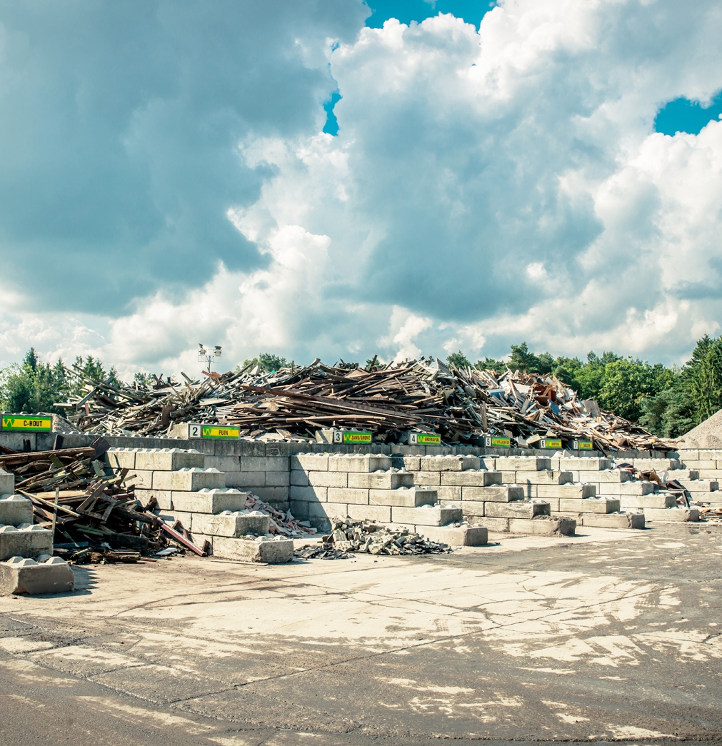 Genummerde opslagvakken van betonblokken op een recyclingvestiging van Van Werven, met borden voor hout, puin en zand en een grote berg sloophout erachter