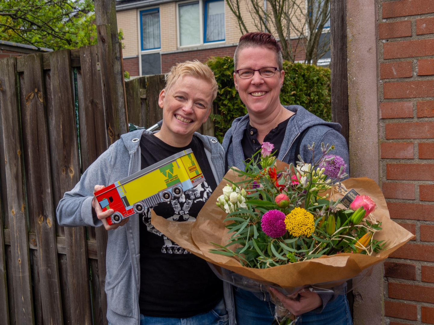 Twee lachende vrouwen met een bloemboeket en een blikken Van Werven-vrachtwagentje voor de schutting van hun woning