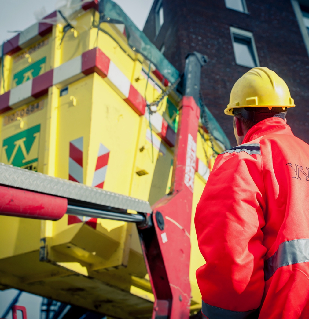 Medewerker met helm en oranje jas kijkt toe hoe een kraan een gele afzetcontainer van Van Werven optilt langs een woonblok