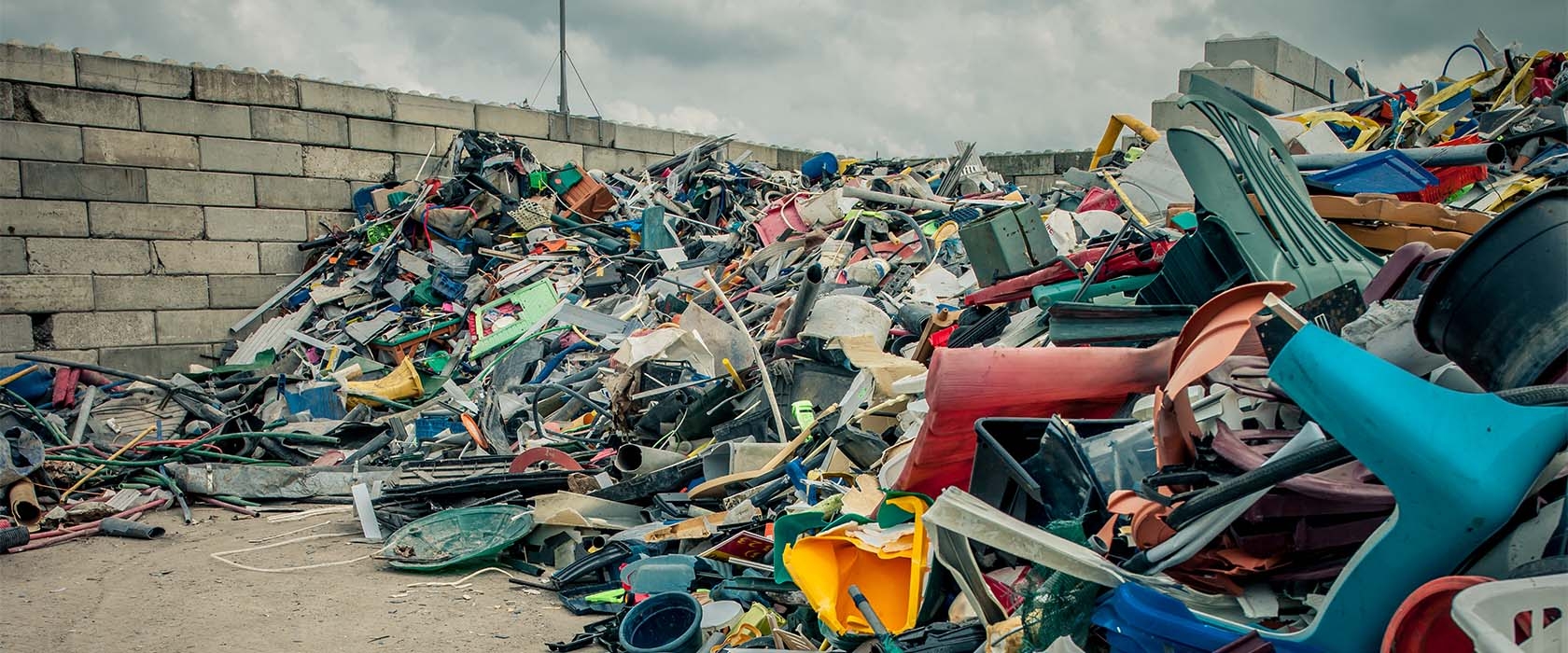 Grote berg gemengd kunststofafval tegen een betonnen keerwand, met tuinstoelen, buizen, emmers en gekleurd plaatmateriaal door elkaar