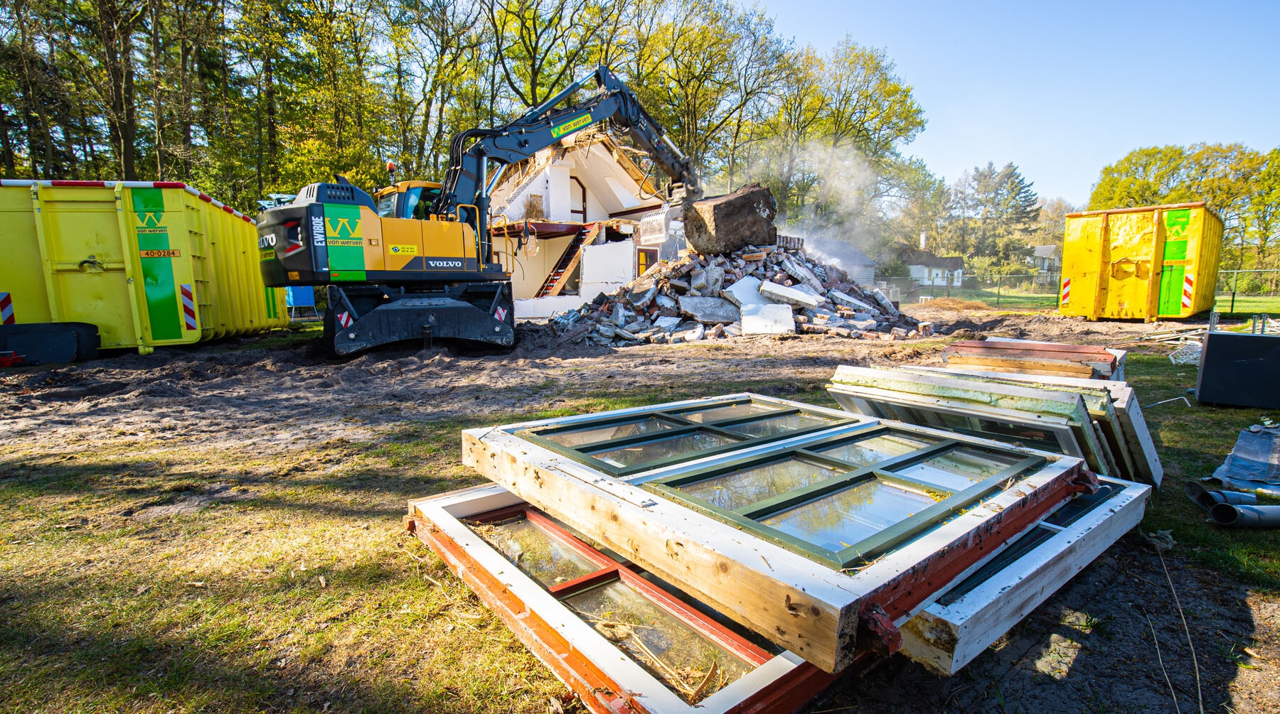 Rupskraan met sloopschaar breekt een betonnen bedrijfspand af, met een stofnevel boven het puin