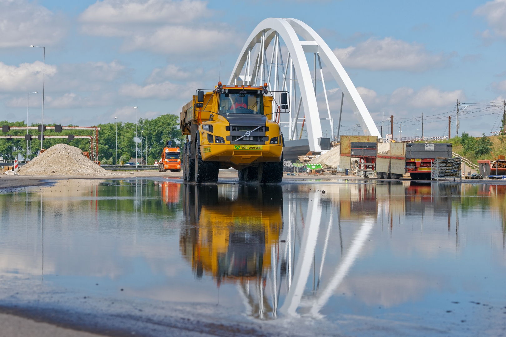 Knikdumper van Van Werven rijdt door een waterplas, met de witte boog van een nieuw viaduct erachter